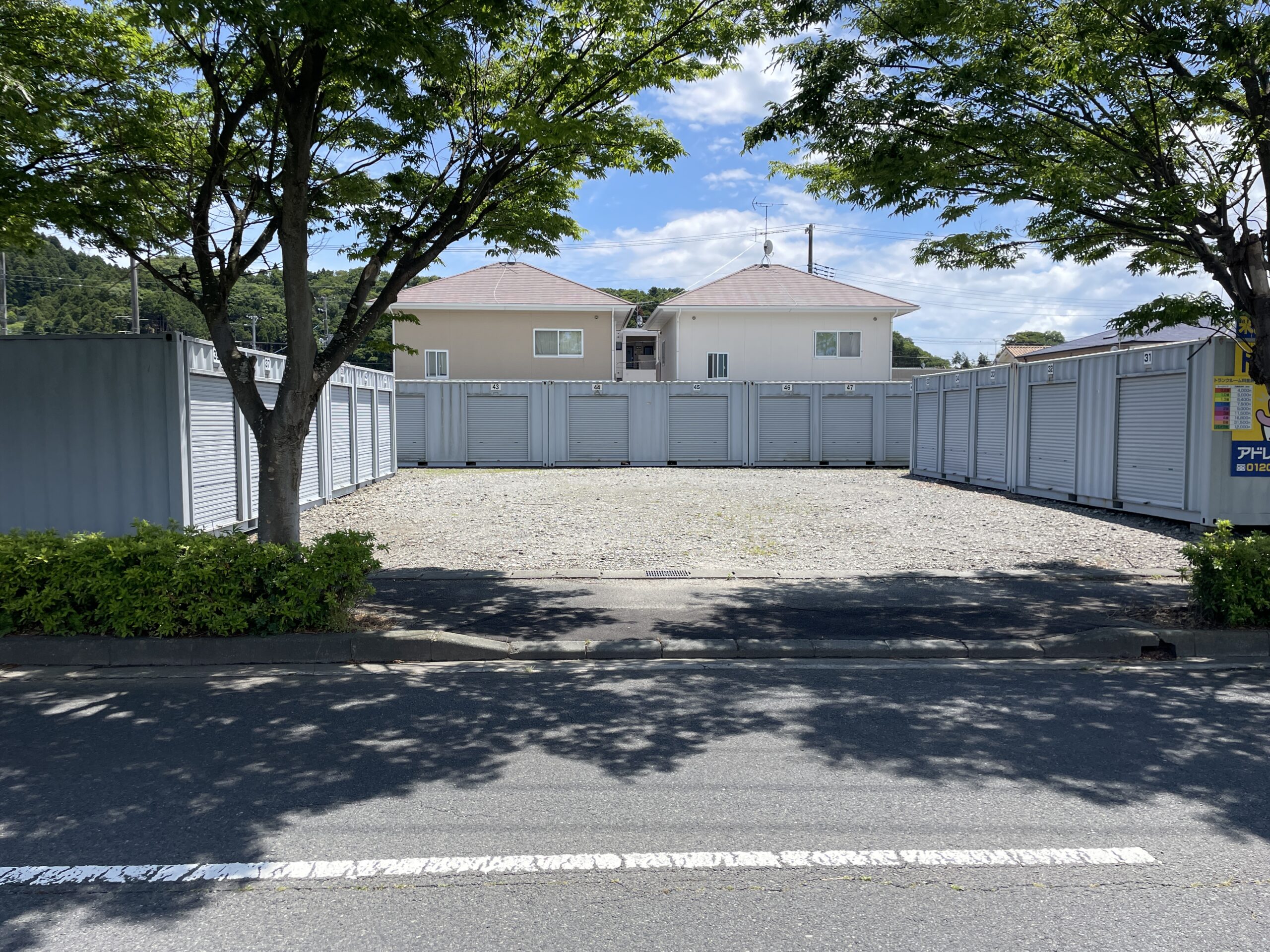 屋外に並んだシルバーのコンテナ型トランクルーム。手前に停まった車のトランクから段ボール箱を運び出す男性の様子。青空の下