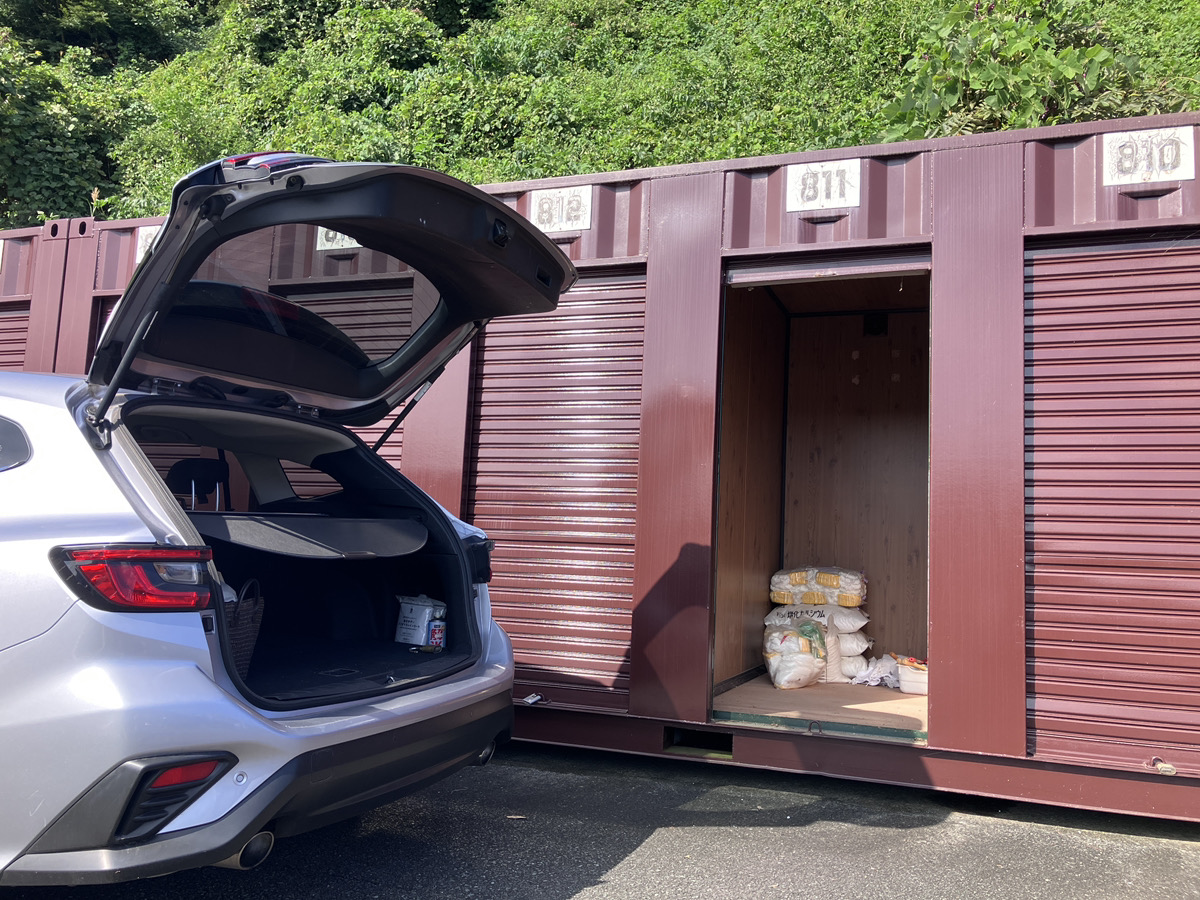 屋外に並んだシルバーのコンテナ型トランクルームの前に車を横づけし、シャッターを開けて段ボールを運び入れる男性