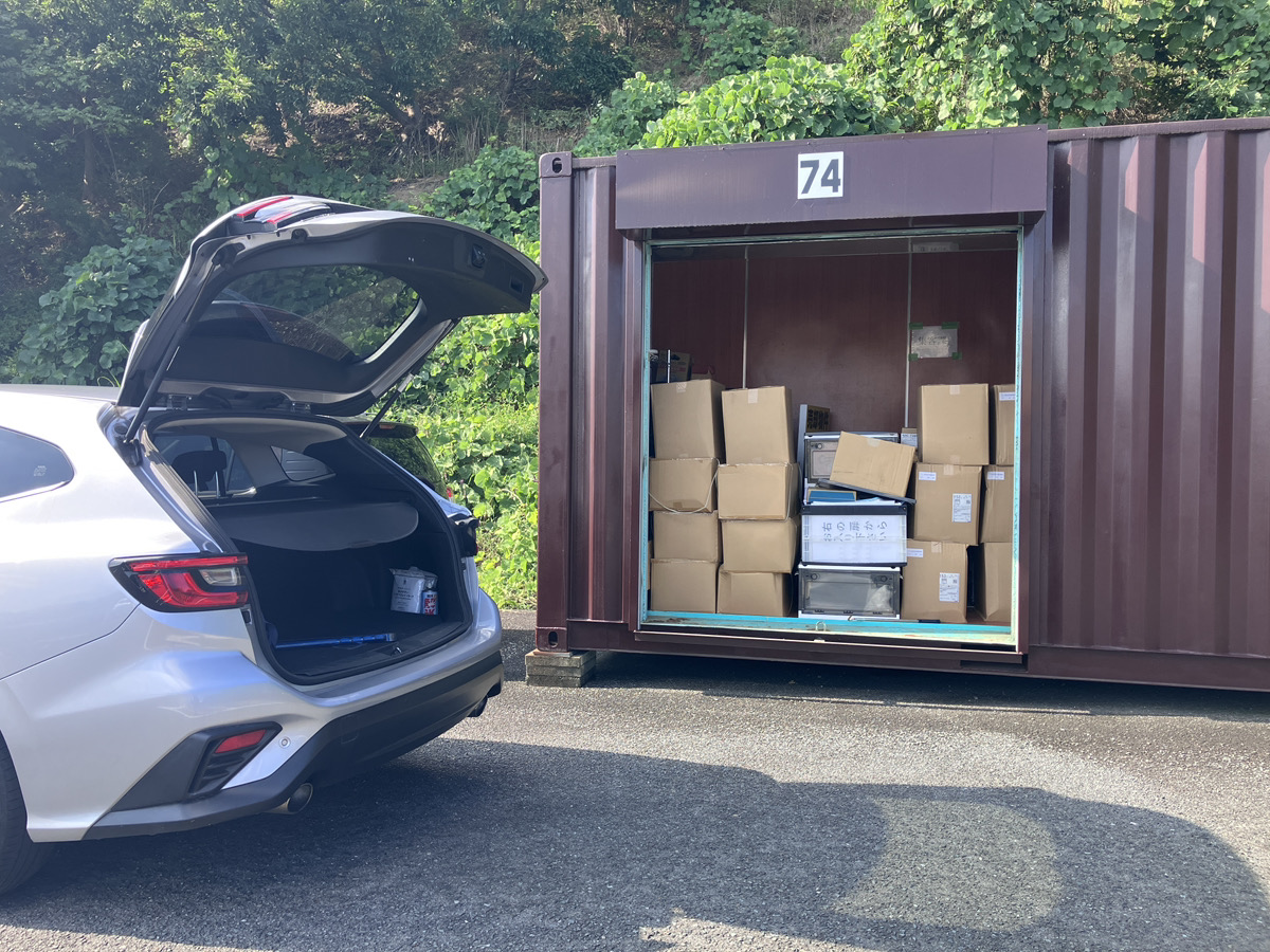 屋外に並んだシルバーのコンテナ型トランクルーム。手前ではシャッターが開けられ、車を横づけしてダイニングテーブルや段ボールを運び込む男性