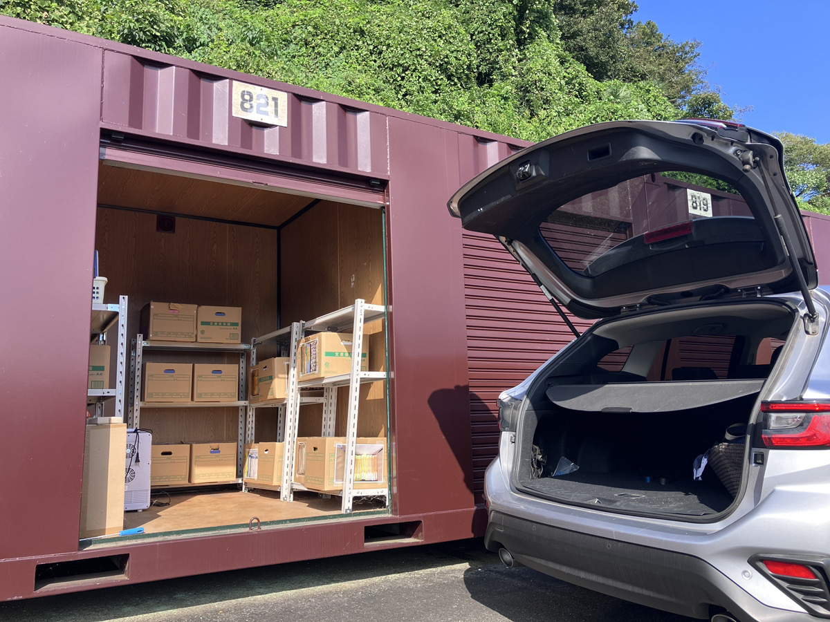 大きな道路沿いに整然と並んだ屋外コンテナ型トランクルーム。手前で車を停めてシャッターを開け、段ボールを運び入れる利用者。