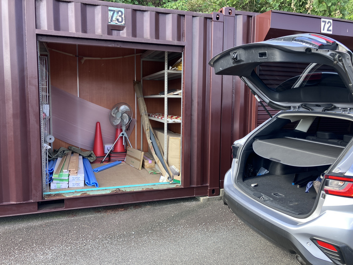 大きな道路沿いに並んだシルバーのコンテナ型トランクルーム。前に車を停めて、シャッターを開けて荷物を運び入れる利用者の様子