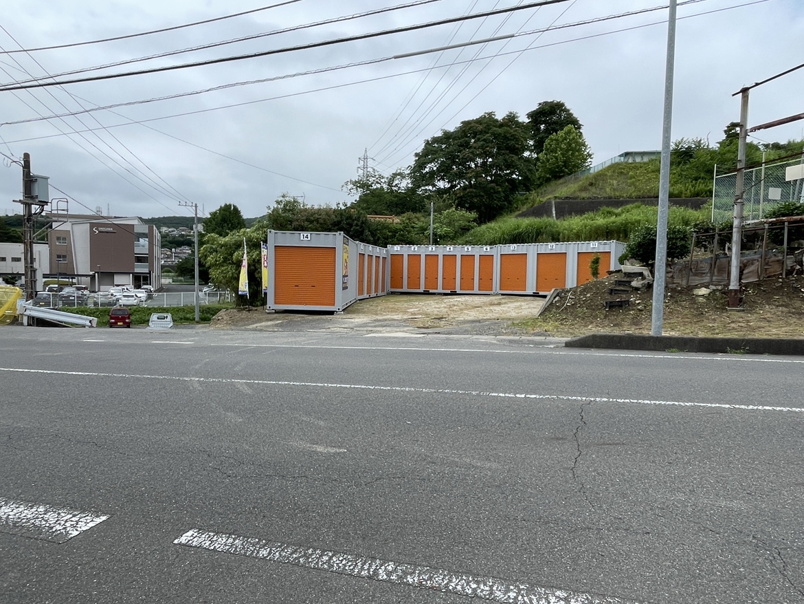 道路沿いに整然と並ぶ屋外コンテナ型トランクルームの前に車を横付けし、シャッターを開けて段ボール箱を運び込む人物