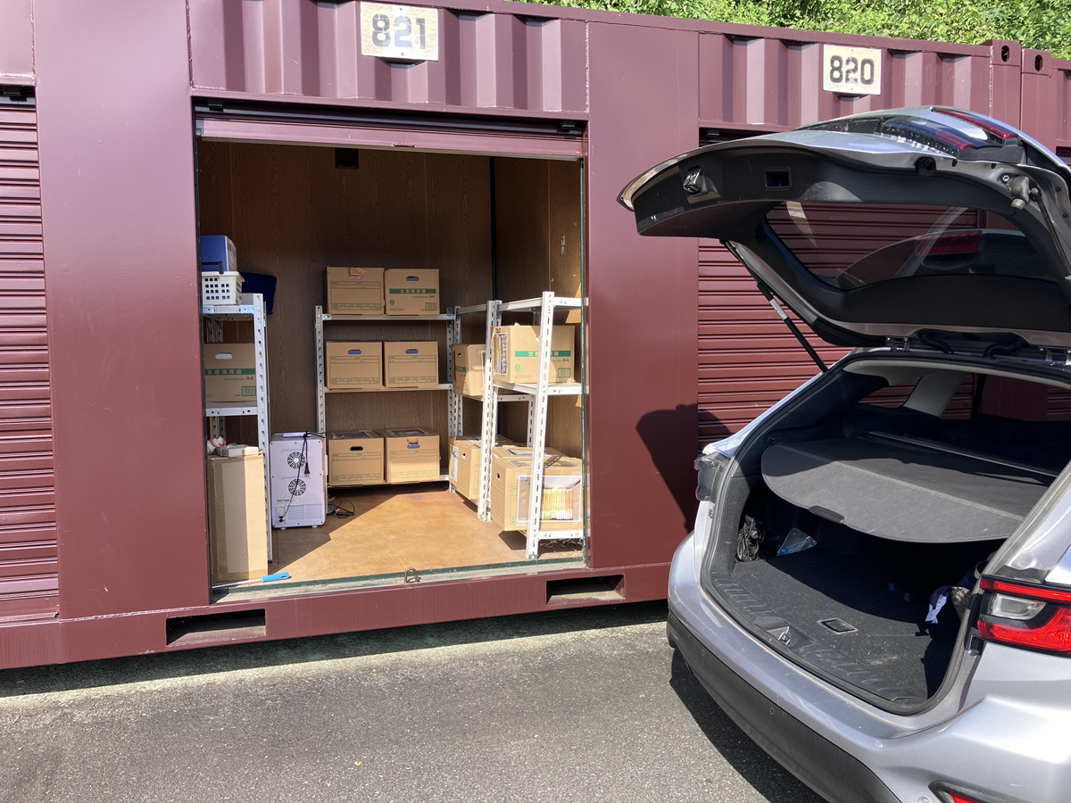 屋外に並ぶシルバーのコンテナ型トランクルームの前に車を横付けして、トランクから電動グリルの箱を運び出す男性