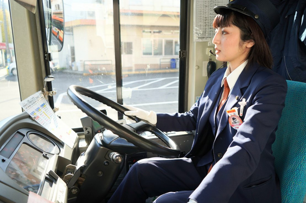 伊豆の温泉街を背景に、笑顔で乗客を迎える女性バス運転士