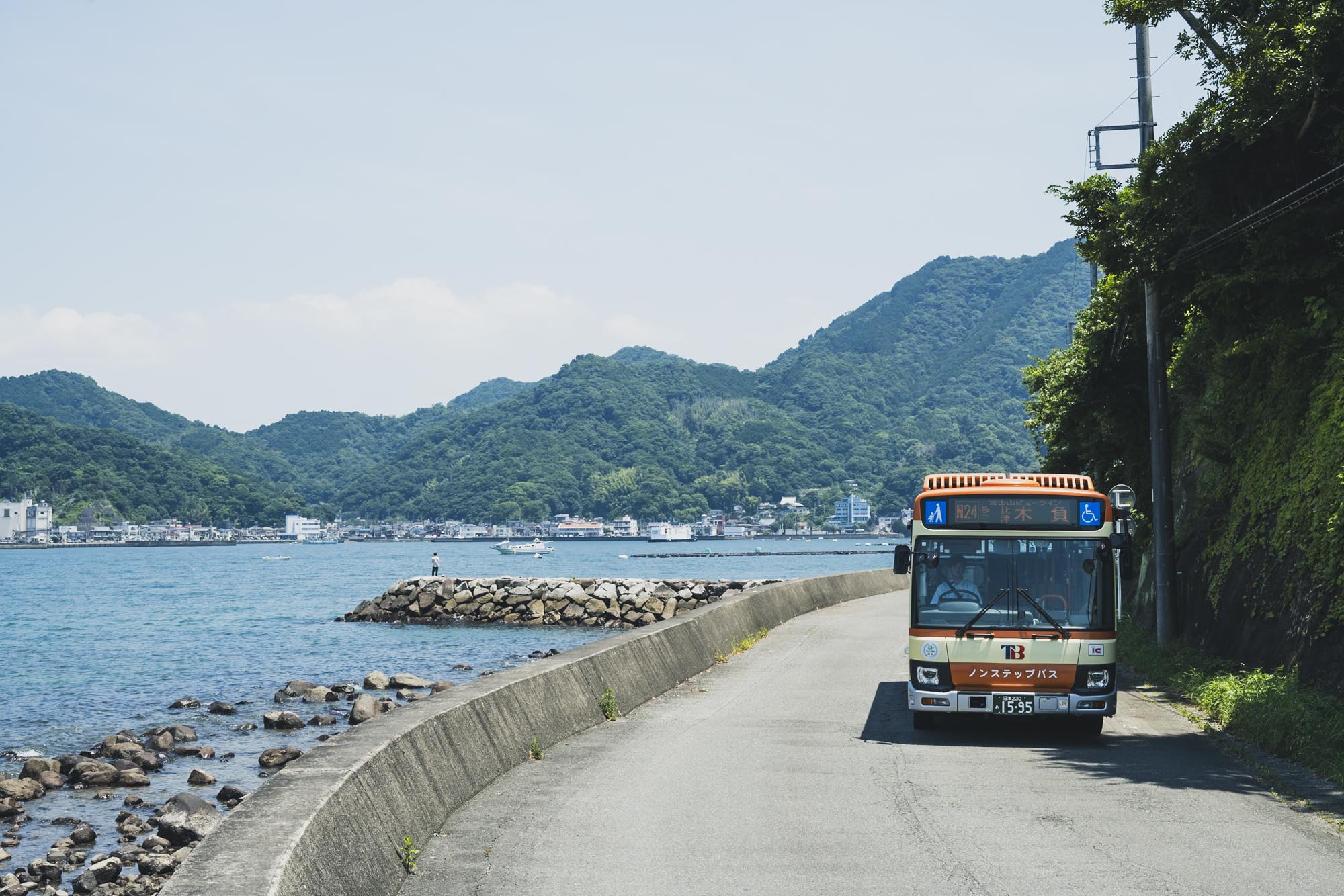 伊豆の海沿いの道を走る路線バスとその前で笑顔で立つ制服姿のバス運転士