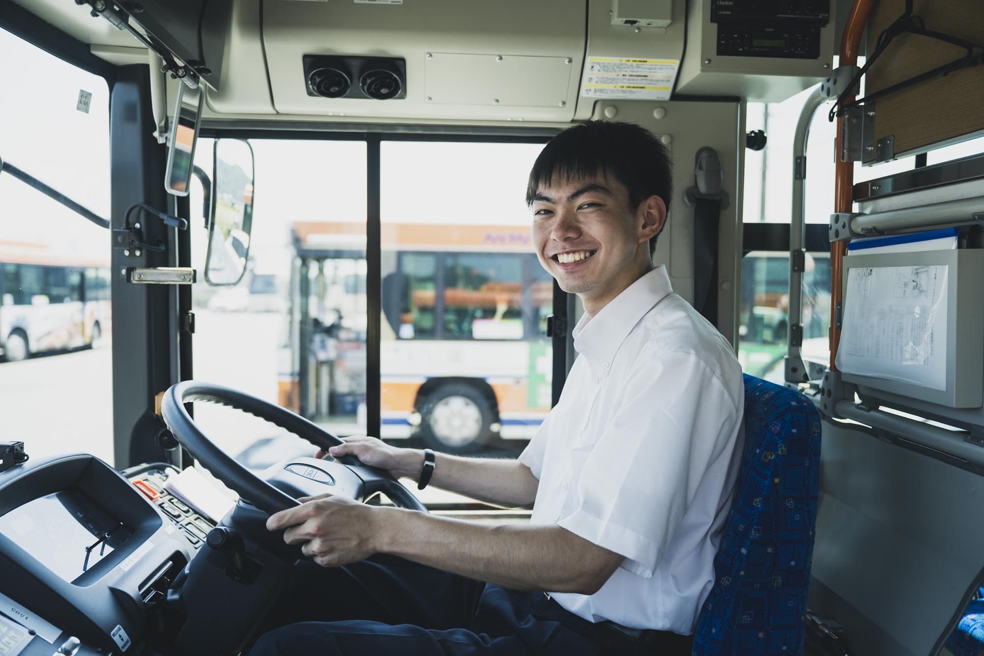 制服姿で笑顔で立つ40代の男性バス運転士、営業所の車庫を背景に安心感のある雰囲気