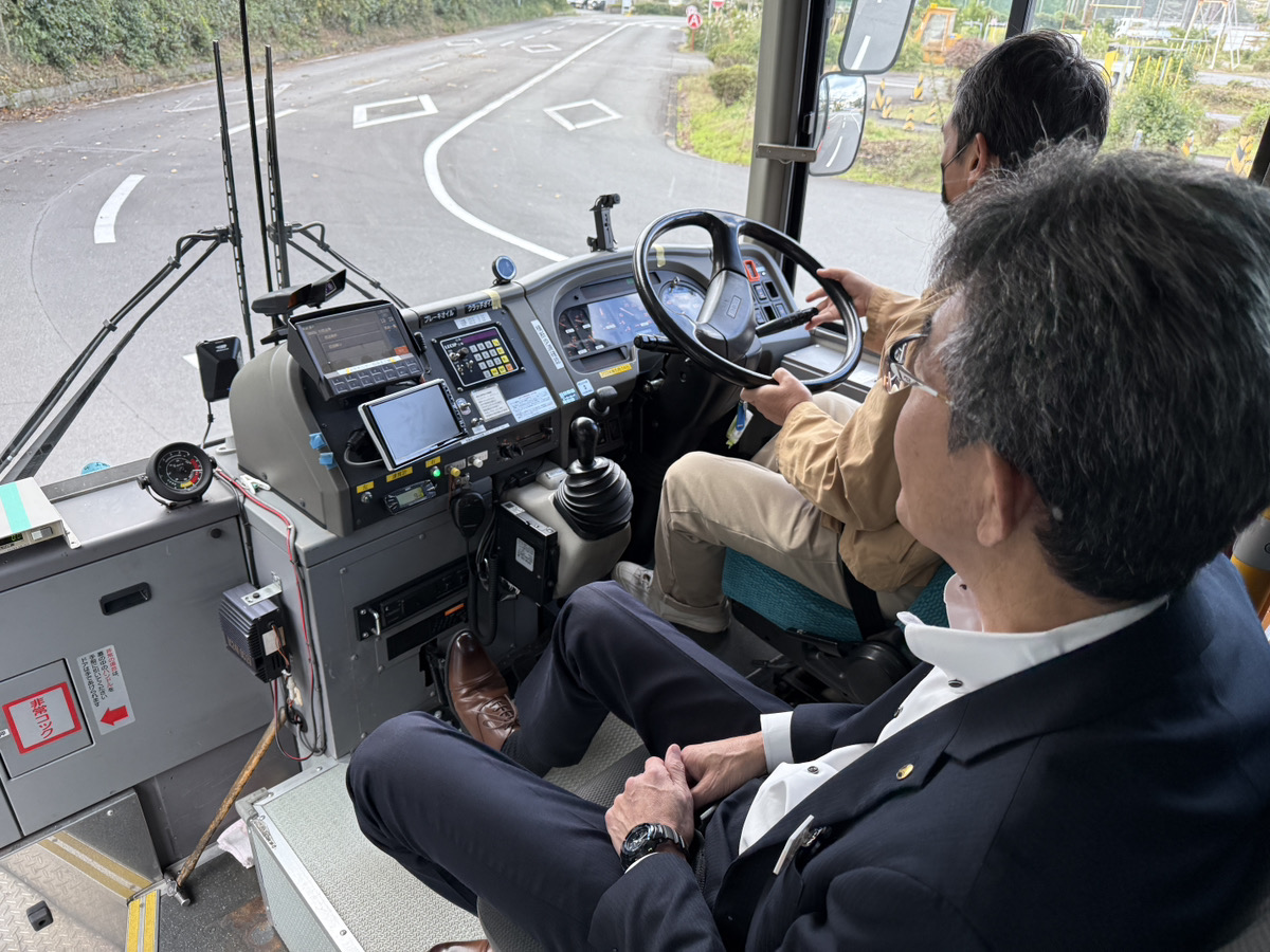 実際のバスの運転席に座り、インストラクターの説明を受けながら初めての操作に驚く笑顔の30代男性
