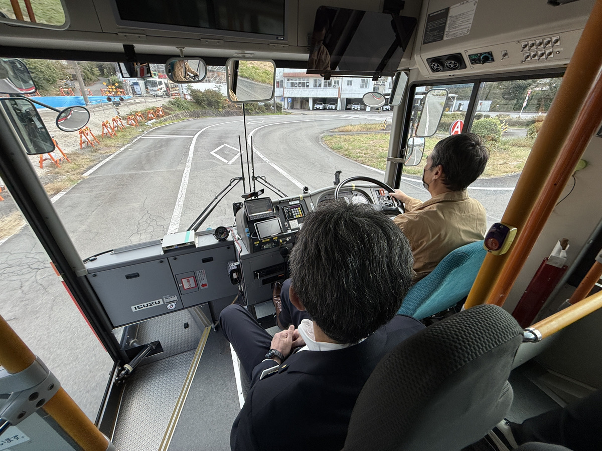 敷地内でバスの運転席に座り、初めての運転に驚きと笑顔を見せる私服姿の30代体験参加者、横で説明する制服姿のスタッフ