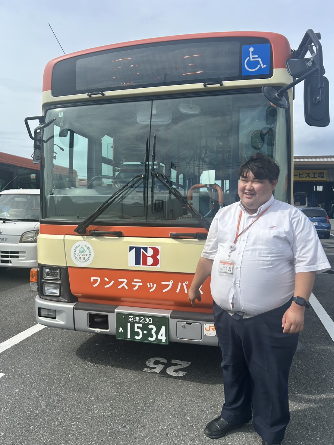 バスの前で立つ運転士の男性。晴天の伊豆の空を背景に、誠実で前向きな表情。全身の立ち姿