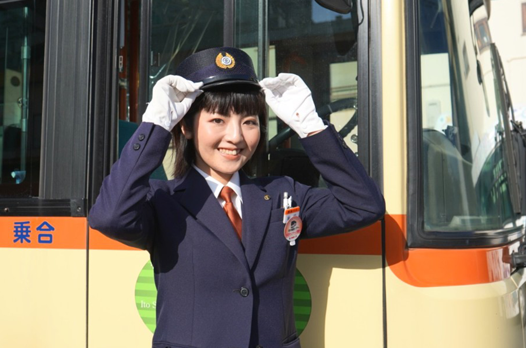 地方の青空の下、路線バスの前に立ち穏やかに微笑む制服姿のバス運転士